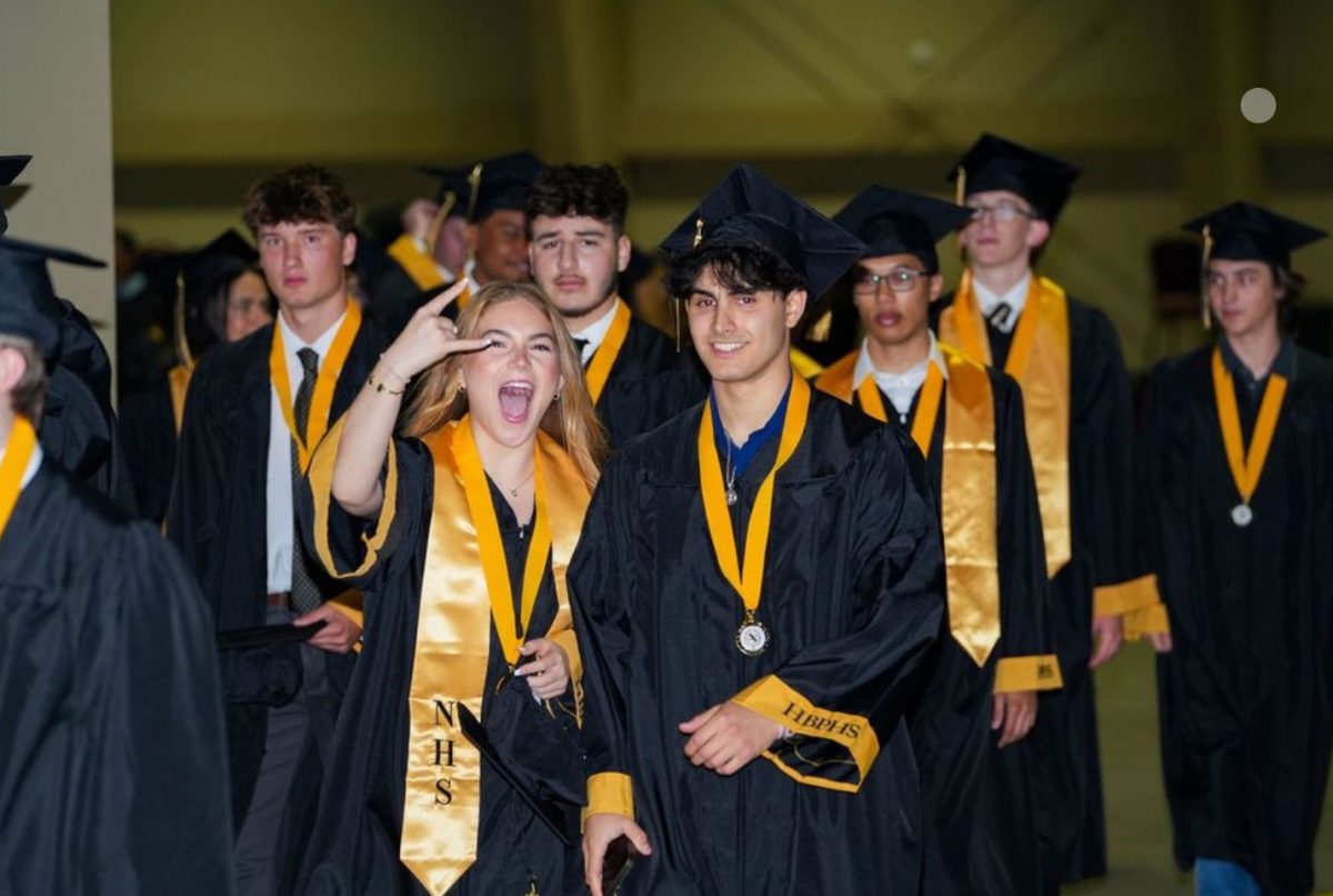 Graduating seniors walk in procession during their commencement ceremony, dressed in black caps and gowns with gold honor stoles and medals. One student flashes a celebratory hand gesture, while others smile as they mark the milestone achievement.