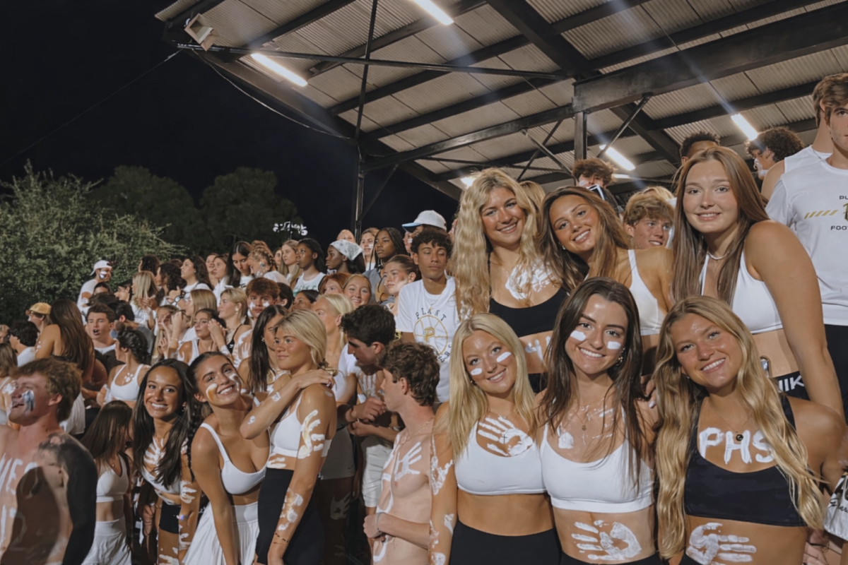 Students from Plants Gold Rush club cheer in the stands during a football game, dressed in white for the theme of the night. The students gather in the “student section” of the bleachers to support their team.  